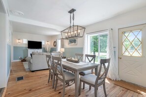 A dining area with a wooden table and chairs, a chandelier, and a television in the background. - Updated, charming home with huge yard, fire pit, & WiFi - walkable location (South Haven)