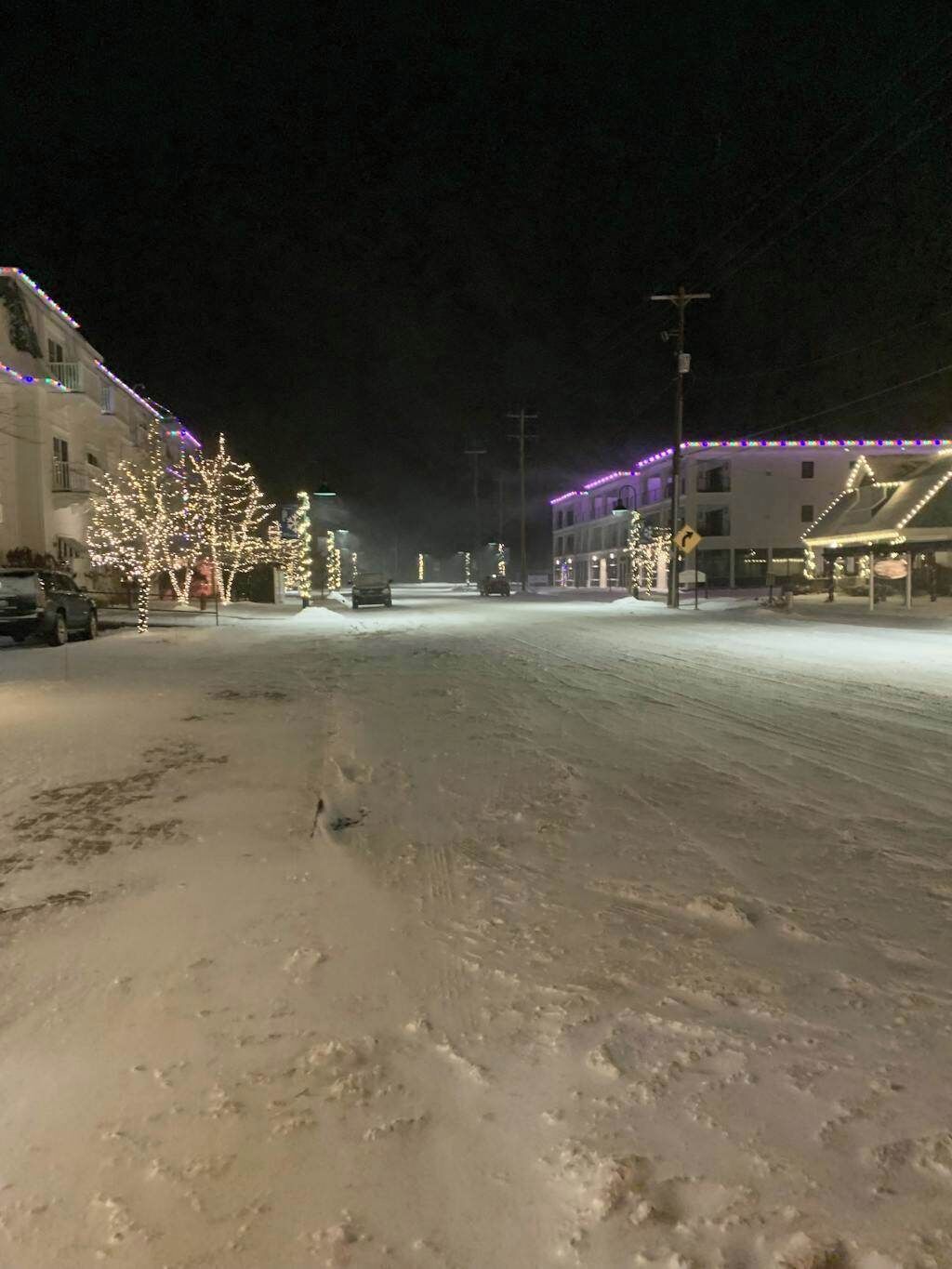 Une rue enneigée, de nuit, bordée de bâtiments des deux côtés, dont certains sont illuminés de décorations de fêtes.