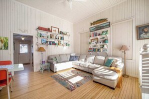 A cozy living room with a white sofa, a small table, and a bookshelf filled with books and decorative items. - Historic cozy cottage in Walloon Lake village close to Boyne Mountain skiing! (Walloon Lake)