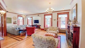 A spacious living room with a sofa set, a red armchair, and patterned armchairs. A fireplace with a mirror and decorative items is visible. - Historical dog-friendly home with fenced yard, grill, Ping-Pong, fireplace, & AC (Manistee)