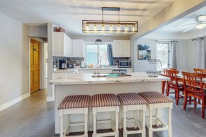 A kitchen with a central island, bar stools, and a dining area with wooden chairs. - Cozy Home Located near Lakes & Trails, large backyard, eV charger & WIFI (Lake Leelanau)
