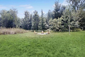 A grassy outdoor area with a fire pit and chairs, surrounded by trees. - Cozy Home Located near Lakes & Trails, large backyard, eV charger & WIFI (Lake Leelanau)