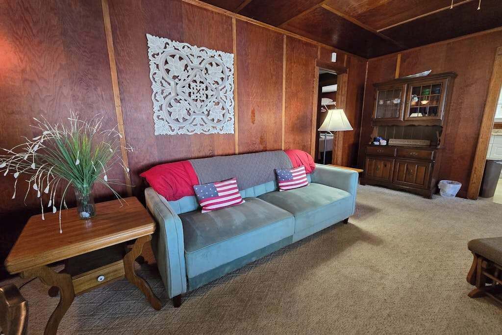 A room with a wooden panel wall, a blue sofa with American flag pillows, a wooden side table with a vase, and a cabinet with glass doors.