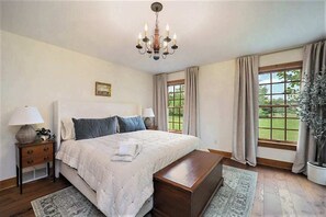 A bedroom with a large bed, a chandelier, a nightstand, a bedside table, a window with curtains, and a view of greenery outside. - Exquisite Cape Cod home with large yard, fireplace, central AC, & W/D (Maple City)