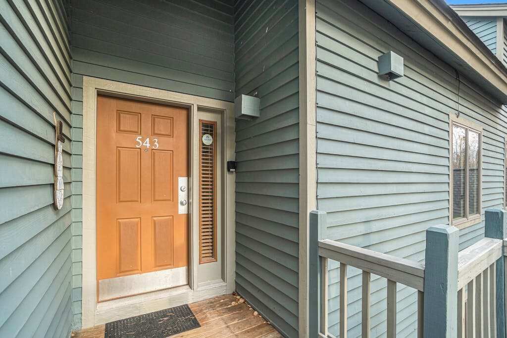 A house with a wooden front door labeled '543,' a small porch with a wooden railing, and a mailbox on the wall.