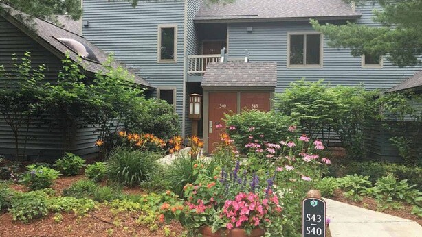 Une maison à deux étages avec un porche à l’avant, une boîte aux lettres et un jardin où poussent différentes plantes et fleurs.