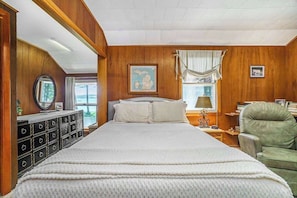 A bedroom with a large bed, wooden paneling, a dresser, a chair, and a window with curtains.