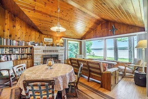 A wooden interior with a fireplace, bookshelves, a dining table, and large windows overlooking a body of water.
