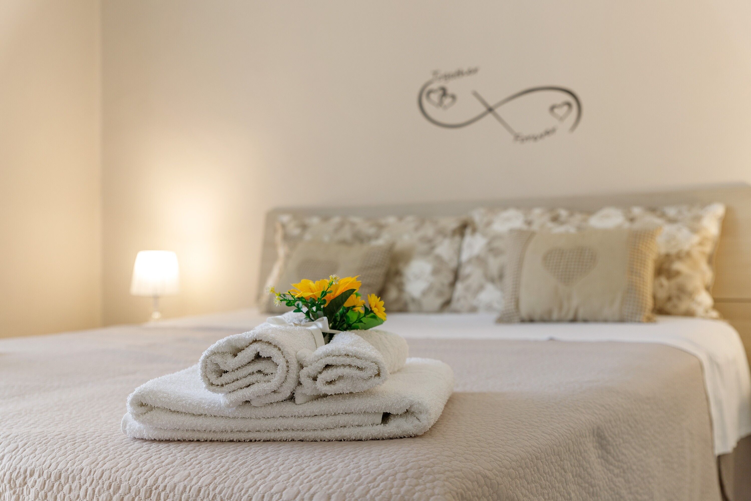 A neatly made bed with rolled towels and a vase of yellow flowers, a wall with a heart and infinity symbol, and a bedside lamp.