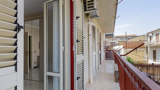 A balcony with a red railing, a sliding glass door, and a view of neighboring buildings. - Apartment "Rina Centro Sicilia" (Pietraperzia)