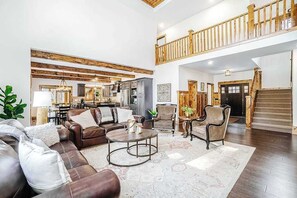 A spacious living area with a high ceiling, wooden beams, a staircase, and a view of the kitchen.
