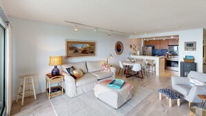 Un salon moderne comprenant un canapé, un pouf et une petite table d’appoint avec une lampe. On y trouve également un coin repas avec une table et des chaises, ainsi qu’une cuisine avec un comptoir et des appareils électroménagers.