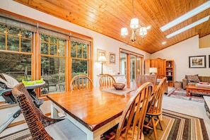 A dining area with a wooden table and chairs, a chandelier, and large windows. - Dog-friendly home with gorgeous garden & decks - stroll to town/the lake (Glen Arbor)