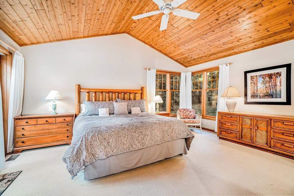 A bedroom with a wooden ceiling, a bed with a patterned bedspread, a nightstand, a chair, a dresser, and a window with curtains.