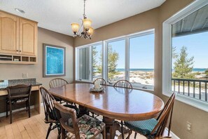 A dining area with a large wooden table, chairs, a chandelier, and large windows with a beach view. - Lakefront condo with deck, gorgeous view, fireplace, & W/D (Glen Arbor)