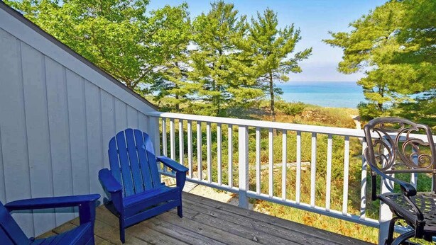 Een houten terras met twee blauwe Adirondack-stoelen en een metalen stoel, met uitzicht op een kustlandschap met bomen en de zee.