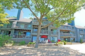A multi-story building with balconies, outdoor seating, and a tree in the foreground. - Beachfront condo with lake views, washer/dryer & fireplace (Glen Arbor)