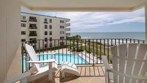 A balcony with a pool, white chairs, and a view of the ocean and a building. - Summer Winds 323 - 2 Bedroom Condo (Indian Beach)