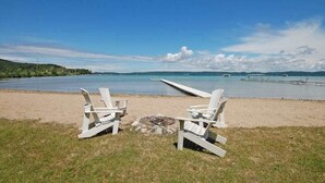 Ein Strand mit zwei weißen Adirondack-Stühlen, einer Feuerstelle und Blick auf das Wasser und das entfernte Land.