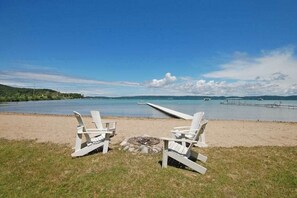 A beach with two white Adirondack chairs, a fire pit, and a view of the water and distant land. - Lakefront cottage with a deck, grill, firepit, dock & partial AC (Glen Arbor)