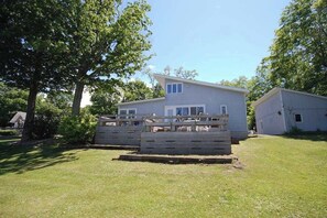 A two-story house with a fenced yard, a wooden deck, and a barn in the background. - Lakefront cottage with a deck, grill, firepit, dock & partial AC (Glen Arbor)