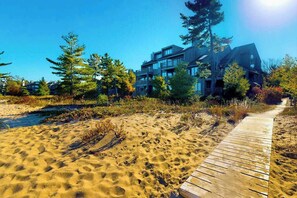 A sandy beach with a wooden boardwalk leading to a modern building with multiple balconies. - Lower-level beachfront condo with deck, fireplace, washer/dryer, central AC (Glen Arbor)