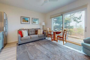 A living room with a sofa, a dining table, chairs, a rug, and a large window with a view. - Desirable beachfront condo in Glen Arbor with patio, fireplace, kitchenette & AC (Glen Arbor)