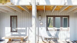 A covered porch with two benches, a wooden ceiling, and a window with a curtain. - Desirable beachfront condo in Glen Arbor with patio, fireplace, kitchenette & AC (Glen Arbor)