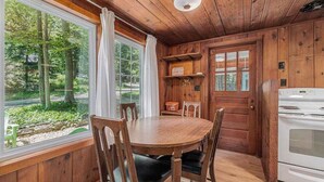 A wooden cabin interior with a dining table, chairs, a white stove, and a window with a view of trees. - Peaceful & shady cottage near beach with streaming, free WiFi, full kitchen (Beulah)