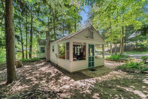 A small, single-story cabin with a porch, surrounded by trees.