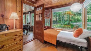 A wooden cabin interior with a bed, a fan, a lamp, and a view of the forest. - Peaceful & shady cottage near beach with streaming, free WiFi, full kitchen (Beulah)