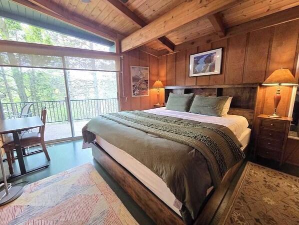 Ein Schlafzimmer einer Holzhütte mit einem großen Bett, zwei Nachttischlampen, einem Esstisch und Blick ins Freie.