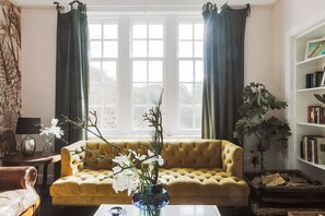Une pièce avec un canapé jaune, une grande fenêtre avec des rideaux, une bibliothèque et une plante.