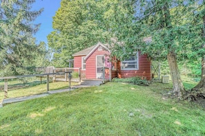 A small red cabin with a wooden deck and a covered porch, surrounded by trees.