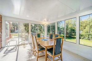 A sunroom with a wooden table and chairs, large windows, and a view of greenery outside.