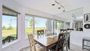 Un coin repas avec une table et des chaises en bois, de grandes fenêtres donnant sur l’extérieur et un miroir au mur.