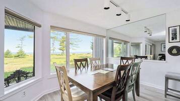 A dining area with a wooden table and chairs, large windows with a view of the outdoors, and a mirror on the wall.