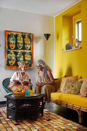 Zwei Personen sitzen auf einem gelben Sofa und einem Korbstuhl mit einer Obstschale.