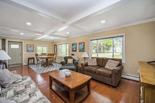 A spacious living room with hardwood floors, a dining area, and a large window offering a view of the outdoors. - Stone home two blocks from the lake & downtown with gas grill & free WiFi (Boyne City)