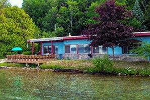 A lakeside cabin with a dock, a covered seating area, and a view of the surrounding forest. - Spacious vacation gem on Crystal Lake w/gas fireplace, WiFi, central AC, firepit (Frankfort)