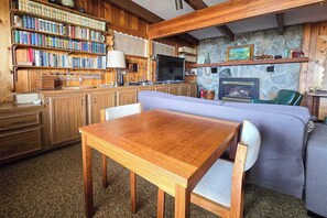 A cozy living room with a wooden table, a sofa, a fireplace, and a bookshelf filled with books. - Spacious vacation gem on Crystal Lake w/gas fireplace, WiFi, central AC, firepit (Frankfort)