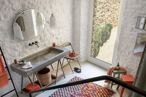 Un baño con lavamanos de piedra, un espejo, luces colgantes, una ventana con vistas y una alfombra estampada.