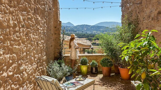 Un patio de paredes de piedra con un banco, plantas en macetas y vistas a colinas distantes.