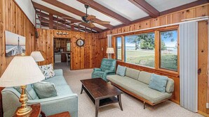 Ein Holzinnenraum mit Sofa, Sessel und Couchtisch, großen Fenstern und einem Deckenventilator.