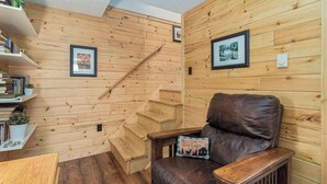L’intérieur d’une cabane avec des murs en bois, un fauteuil en cuir marron, un escalier en bois et une bibliothèque contenant des livres et des objets décoratifs.