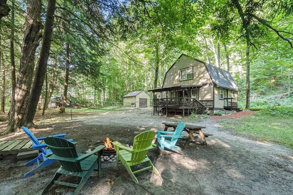 Une cabane dotée d’une terrasse, entourée d’arbres, et un foyer avec des chaises.