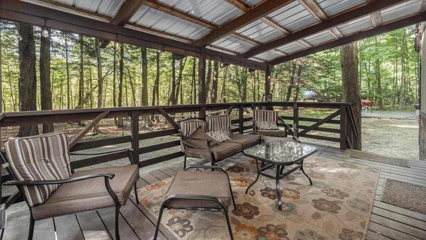 Un espace extérieur aménagé, abrité et doté de chaises à structure métallique et d’une table en verre, entouré d’une terrasse en bois et surmonté d’un toit en métal, offrant une vue sur une zone boisée.