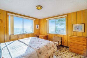 Ein Zimmer mit Holzwänden, einem Bett mit floralem Überwurf, einem Schreibtisch, einem Stuhl und einer Kommode.