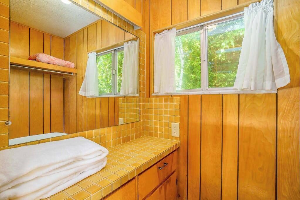 A wooden cabin-style bathroom with a tiled countertop, a mirror, a window with curtains, and a shelf with a towel.