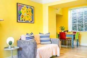 A bright yellow room with a grey sofa, striped pillows, a round table with a lamp, and a window with a stone wall view.